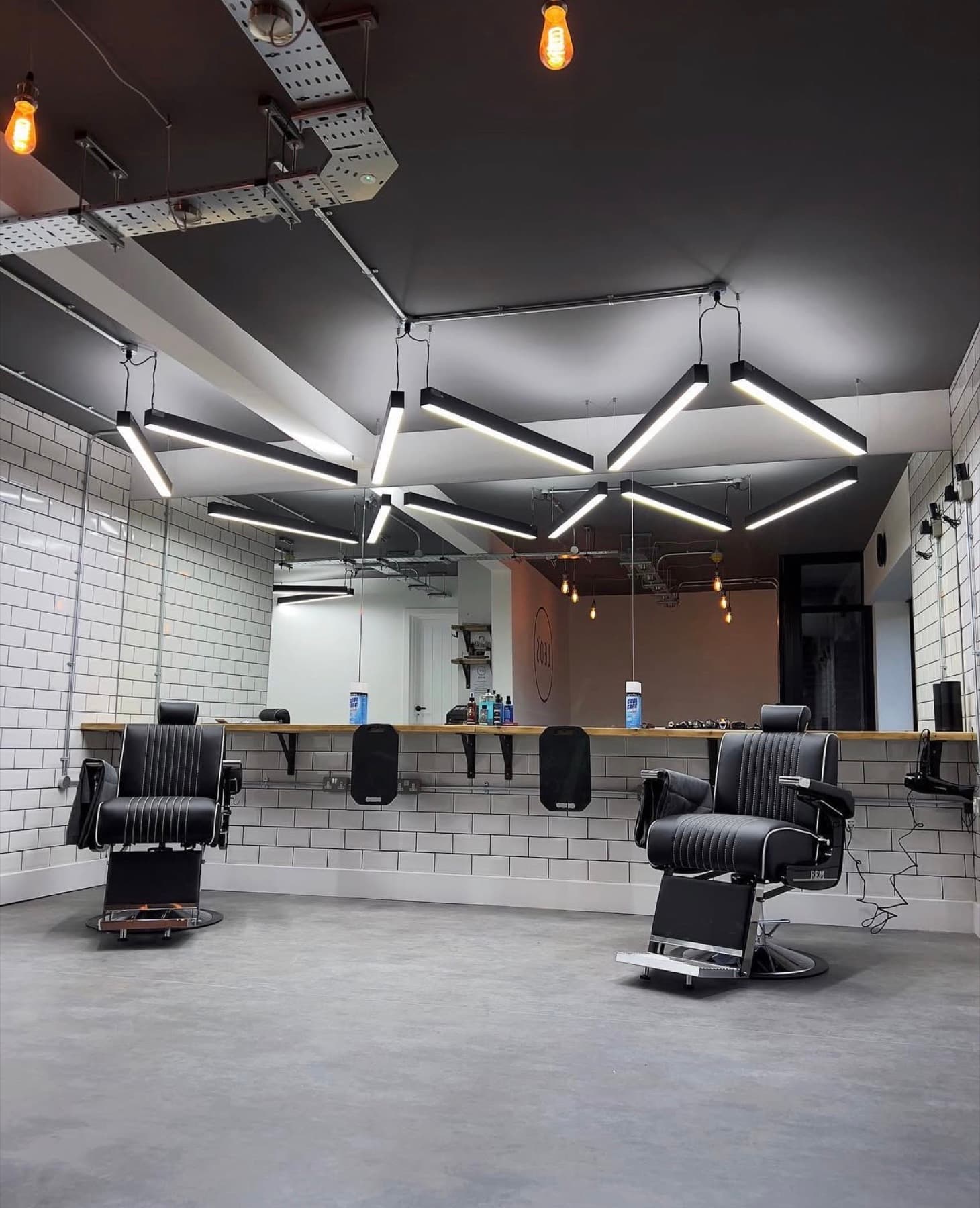 The shop floor at Leo's Barbers, with two black barber chairs on polished concrete, white subway tile and angular LED light bars overhead